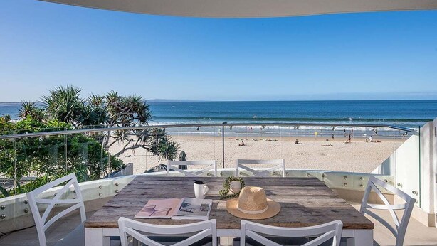 Ein Balkon mit Blick auf den Strand, weiße Stühle, ein Holztisch und ein Hut.