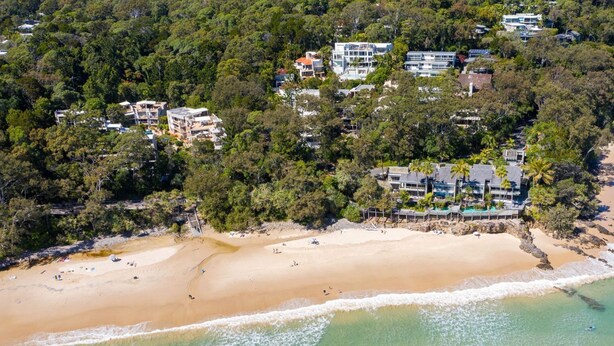 An aerial view of a beach with several buildings, including a large resort with multiple structures and a smaller building with a red roof.