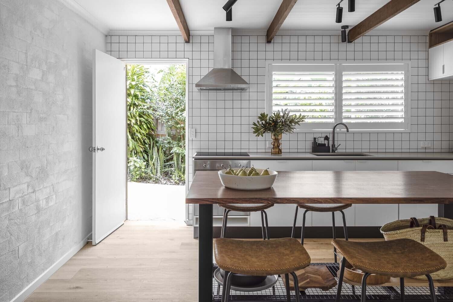 Een moderne keuken met een houten kookeiland, twee barkrukken, een schaal met fruit en een raam met jaloezieën.