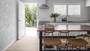 A modern kitchen with a wooden island, two bar stools, a bowl of fruit, and a window with blinds.