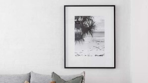 A modern living room with a grey sofa, a framed black and white beach photograph, and a small wooden stool.