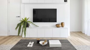 A modern living room with a flat-screen TV mounted on a white wall, a wooden coffee table with a book and decorative items, and a potted plant.
