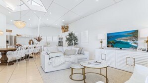 A modern living room with a white sofa, a dining area with white chairs, and a kitchen in the background.