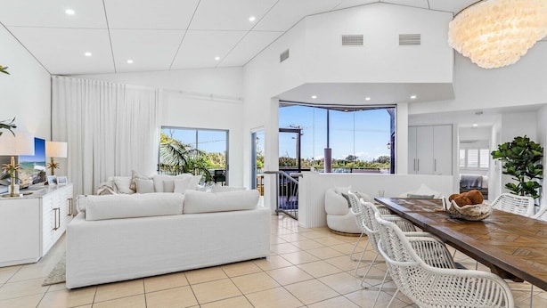 A modern living room with a white sofa, dining area with white chairs, and large windows offering a view of greenery.