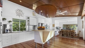 A modern kitchen with white cabinets, a wooden dining table, and a large window overlooking greenery.
