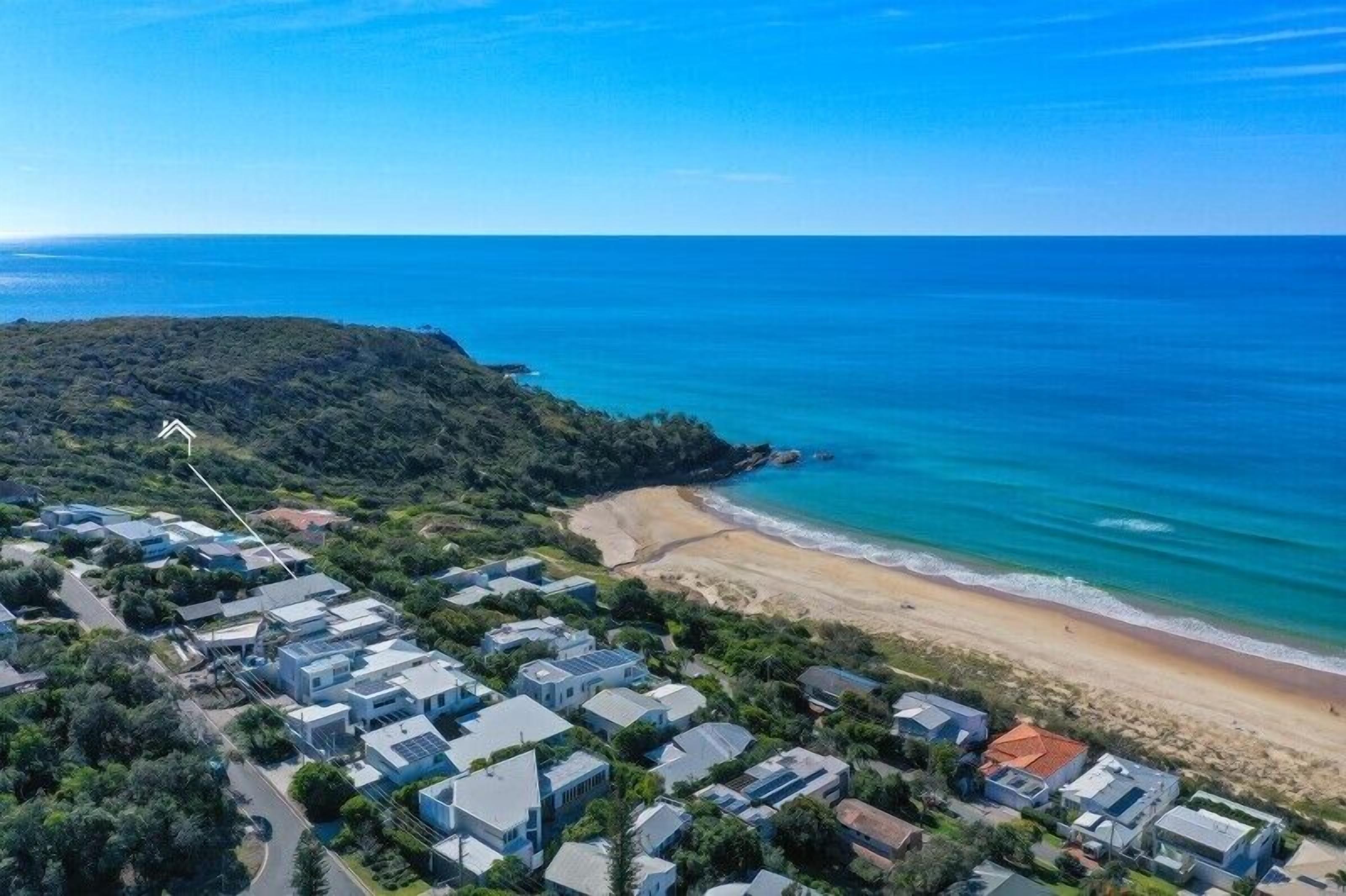 Vue aérienne d’une zone résidentielle côtière, avec des maisons, une plage et une colline boisée.