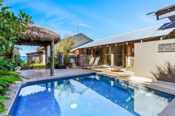 Ein Swimmingpool mit einer strohgedeckten Konstruktion, einer überdachten Terrasse und einem Gebäude mit einer Metallbedachung.