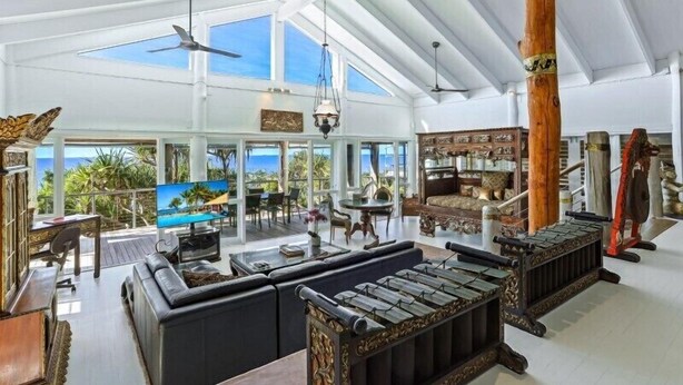 Un vaste espace de vie agrémenté d’une grande fenêtre donnant sur un paysage tropical, d’un coin salon confortable et d’un meuble en bois contenant des objets décoratifs.