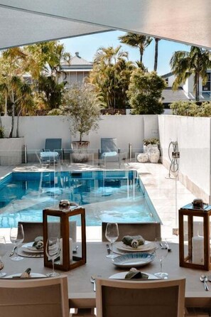 Un espace de restauration aménagé au bord de la piscine, offrant une vue dégagée sur celle-ci, entouré de palmiers et avec un bâtiment blanc en arrière-plan.