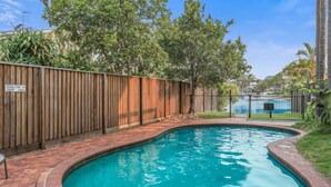 A swimming pool with a wooden fence and a brick border, surrounded by trees and a clear sky. - Riverside location, Noosa Sound (Noosa Heads)