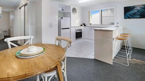 A modern kitchen with white cabinets, a refrigerator, stove, and a sink. There is a dining area with a wooden table and chairs. - Riverside location, Noosa Sound (Noosa Heads)
