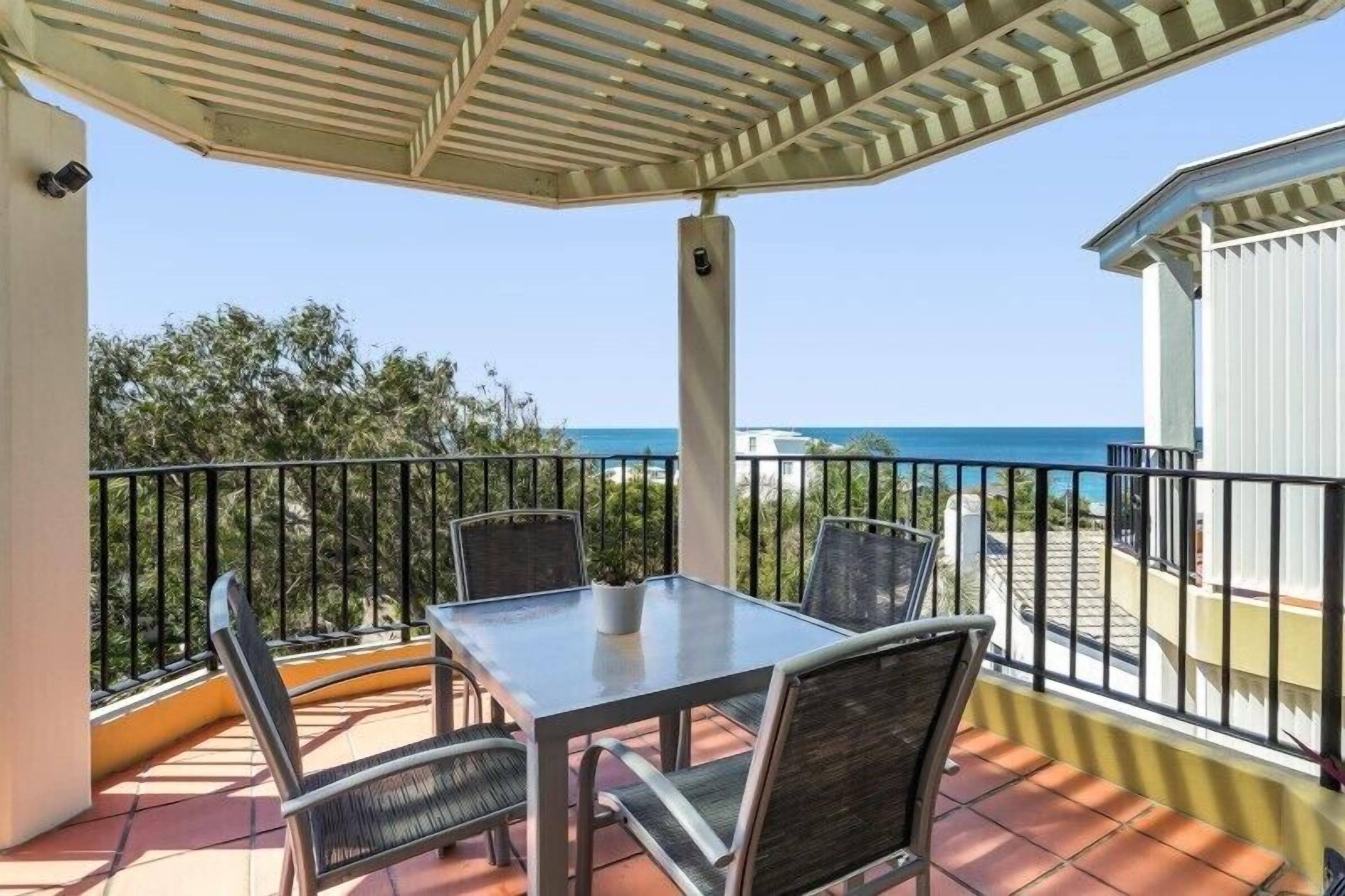 A balcony with a table and chairs, overlooking a coastal view.