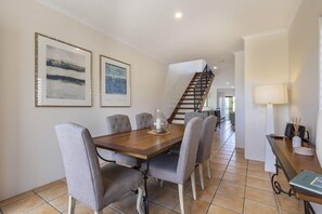 A dining area with a wooden table and six chairs, a staircase in the background, and two framed artworks on the wall. - Wake up to ocean views in stylish comfort, Sunshine Beach (Sunshine Beach)