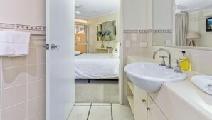 Ein Hotelbad mit einem großen Waschbecken, einem Spiegel und einem durch eine offene Tür erkennbaren Bett.