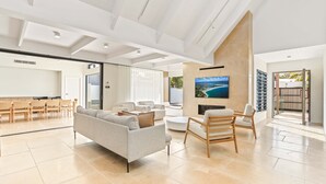 A spacious living room with a large flat-screen TV, a sofa, and a coffee table. There are wooden chairs and a dining area with a table and chairs.