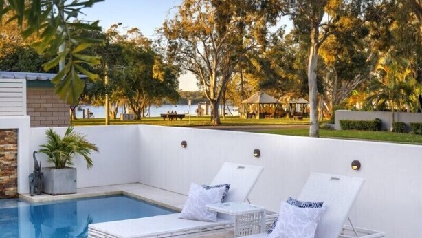 A poolside area with lounge chairs and a small table, surrounded by trees and a view of a park. - Exquisite Riverside Living, Noosaville (Noosaville)