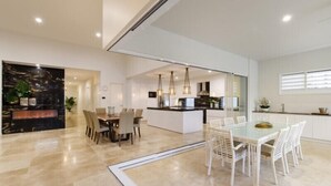 Une cuisine moderne dotée d’un îlot central, de luminaires suspendus et d’un coin repas avec une table en verre et des chaises blanches.
