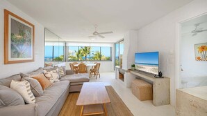 Un salon spacieux avec un grand canapé, une table basse en bois et un téléviseur. Il y a un balcon avec vue sur la plage et les palmiers.