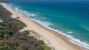 Vue aérienne d’une plage aux eaux d’un bleu limpide, bordée d’un rivage sablonneux et parsemée de quelques plantes.