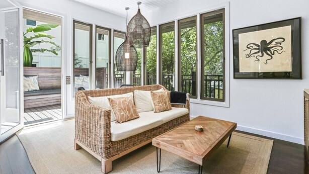 A spacious room with a wicker sofa, a wooden coffee table, and large windows offering a view of greenery outside.
