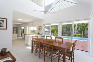 A modern kitchen with a central island, a dining area with a wooden table and chairs, and a view of a swimming pool through large sliding glass doors.