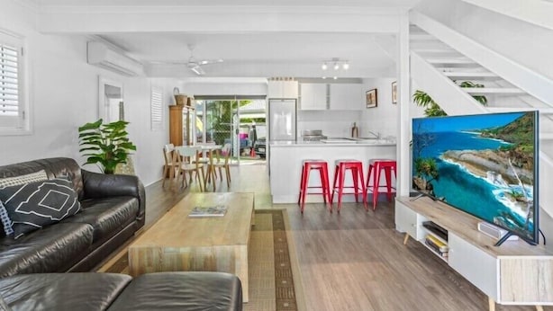 Un salon moderne avec un canapé en cuir noir, une table basse en bois et un téléviseur posé sur un meuble blanc. On y trouve également un coin cuisine avec des placards blancs et des tabourets rouges.