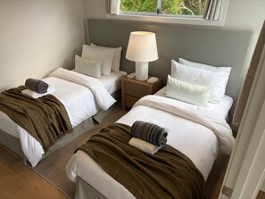 Habitación de hotel con dos camas, una mesita de noche con lámpara y vistas al exterior con vegetación.