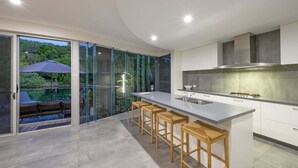A modern kitchen with a large island, bar stools, and a view of a pool through glass doors. - 31 Witta Circle, Noosa Heads (Noosa Heads)