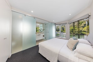 Relax in a serene bedroom with a large bed and sliding door to a bathroom, offering a view of a lush garden and a body of water.