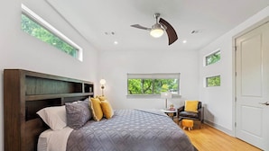 A bedroom with a large bed, a ceiling fan, a window with a view of greenery, a chair, and a door.