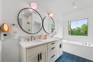 Un baño moderno con dos espejos grandes, un tocador blanco, una bañera y un ventanal con vistas al follaje.