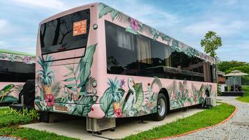 A pink bus with tropical plant and bird decals parked on a gravel path.