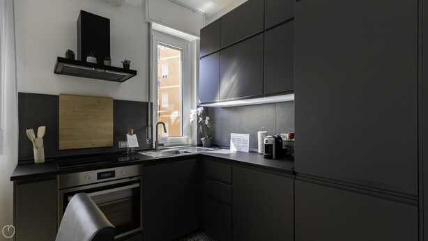 A modern kitchen with dark cabinetry, a stainless steel oven, a wooden board, and a window with a view of buildings. - Italianway Easy - Favretto 13 (Milan)