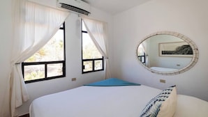 A bedroom with a bed, a window with curtains, an air conditioning unit, and a round mirror on the wall.