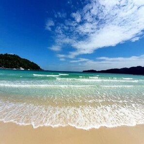 A beach with clear turquoise water, gentle waves, and a distant green hill.