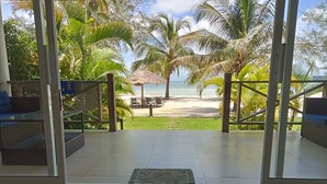 A view from a room with a glass door leading to a beach area with palm trees and a thatched-roof structure. - Saracen Bay Resort (Koh Rong Sanloem)