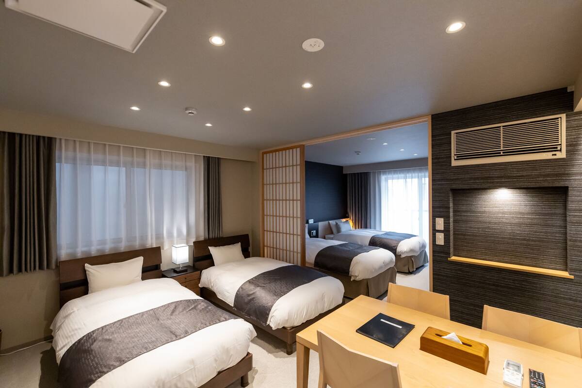 A hotel room with two beds, a desk, and a view of the adjacent room.