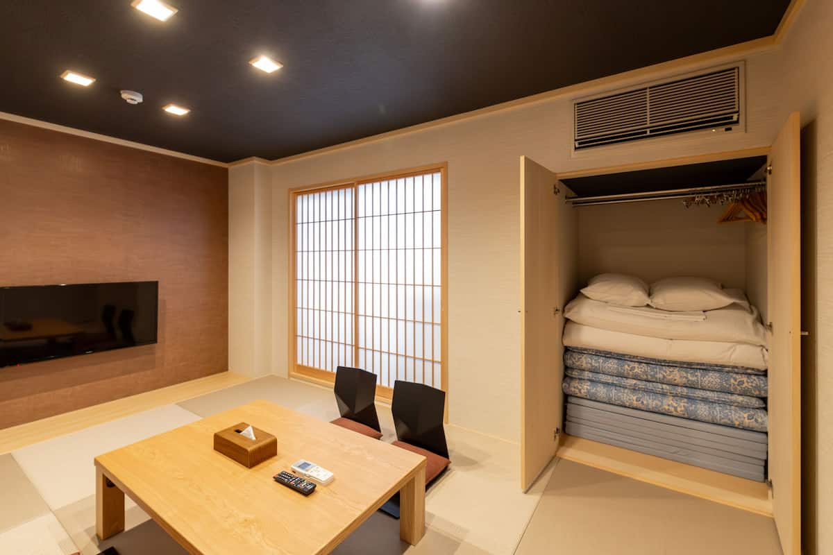 A compact room with a wooden table, two chairs, a TV mounted on the wall, a sliding closet with bedding, and a large shoji screen window.