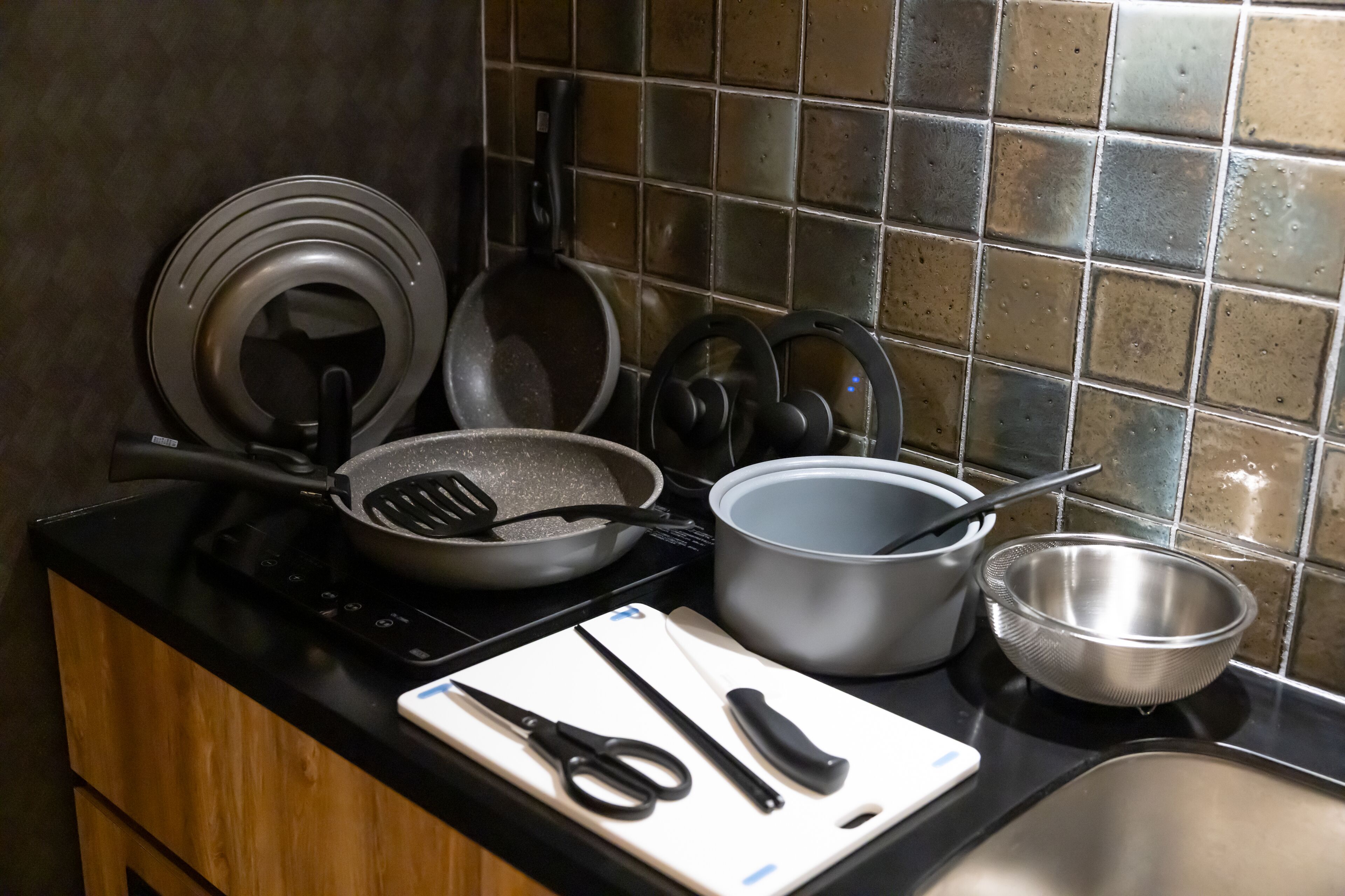 Una cocina con estufa, sartén, olla y tabla de cortar con utensilios.