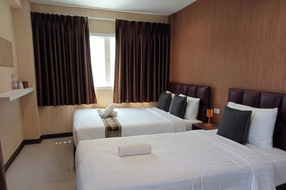 A hotel room with two beds, a wooden headboard, a bedside table with a lamp, and a window with curtains.