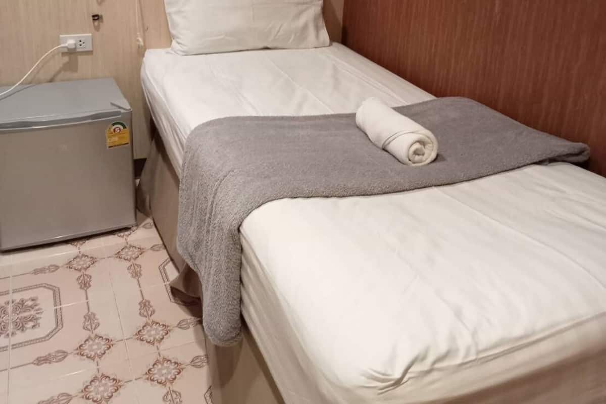 A single bed with a grey blanket and a rolled towel, a small heater, and a power outlet on the wall.