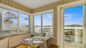 A dining area with a table set for four, large windows with a view of a dock and trees, and a wooden deck outside. - Beach Pass-Charming Waterfront Cottage in Wareham (Wareham)