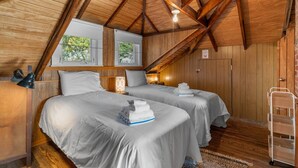 A room with wooden beams, two beds with white linens, a nightstand, a lamp, and a window with a view of trees. - Beach Pass-Charming Waterfront Cottage in Wareham (Wareham)