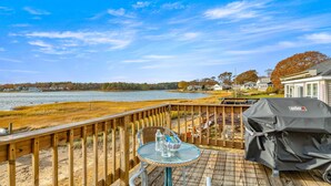 A wooden deck with a glass table and chairs, overlooking a lake with houses in the background. - Beach Pass-Charming Waterfront Cottage in Wareham (Wareham)