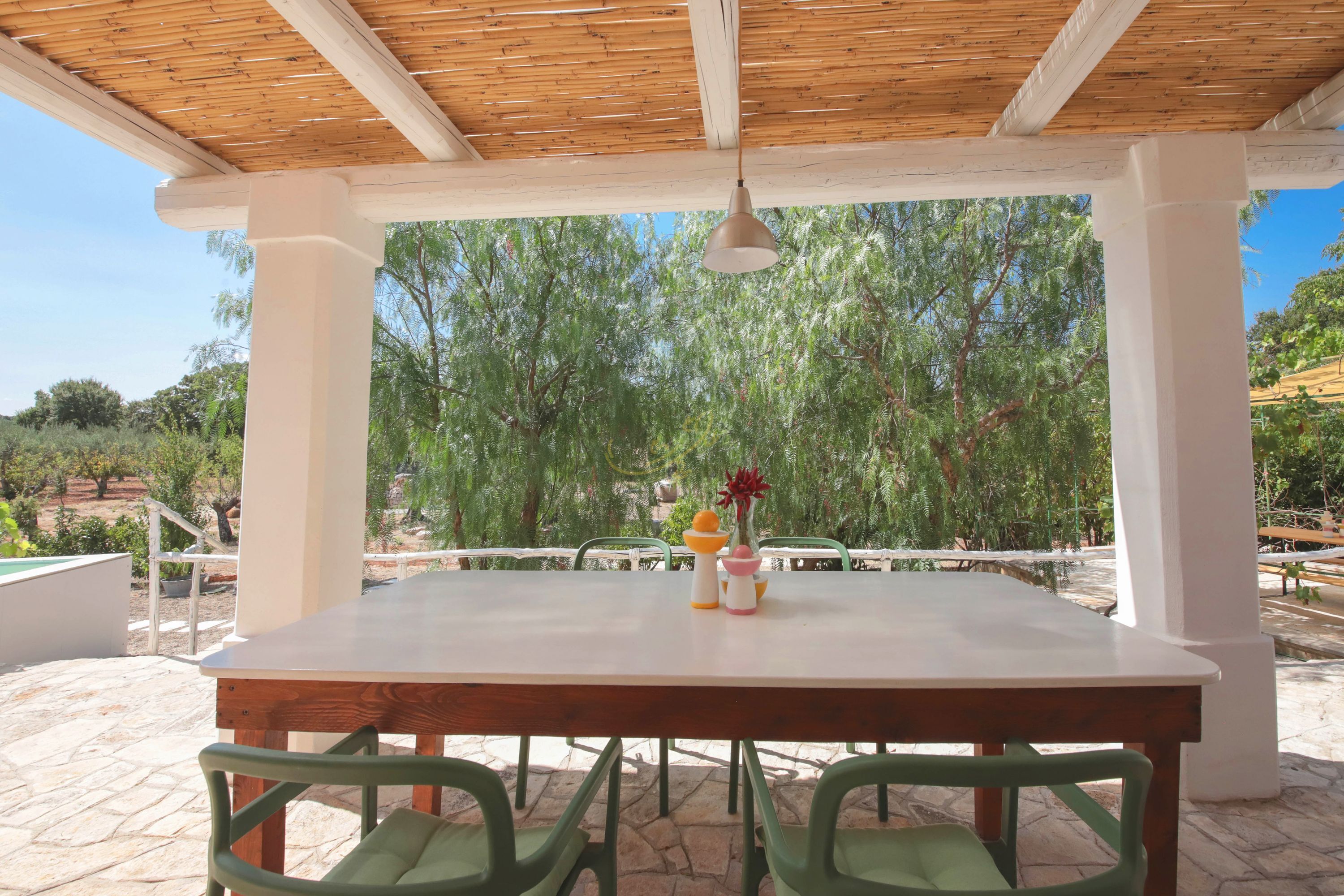 A covered outdoor dining area with a table and chairs, surrounded by greenery and a clear sky.