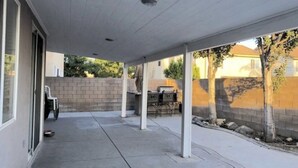 A covered patio area with a concrete floor, white pillars, and a brick wall in the background. - One Bedroom With its Full Bathroom (Fontana)