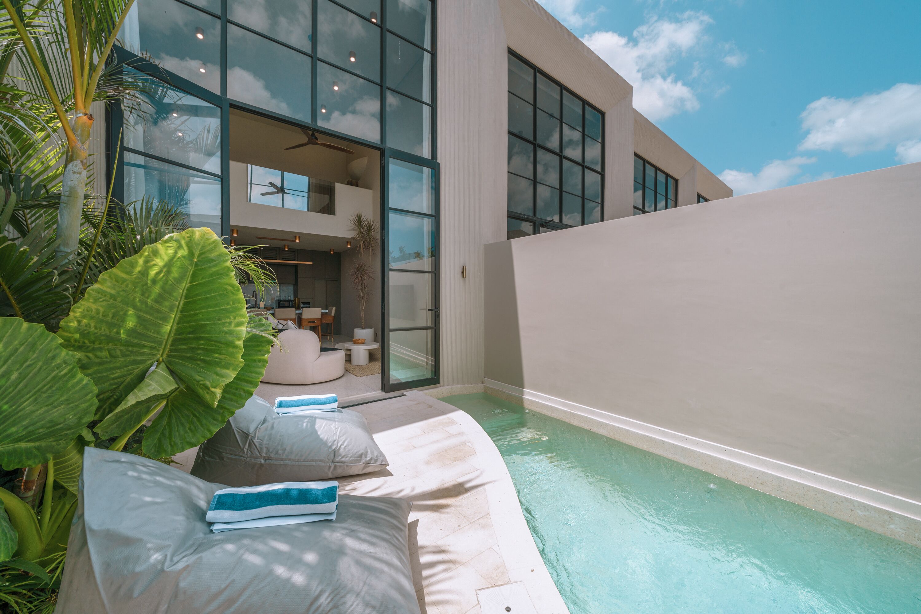 Un edificio moderno con amplios ventanales, una zona de piscina con sillones de descanso y plantas tropicales.