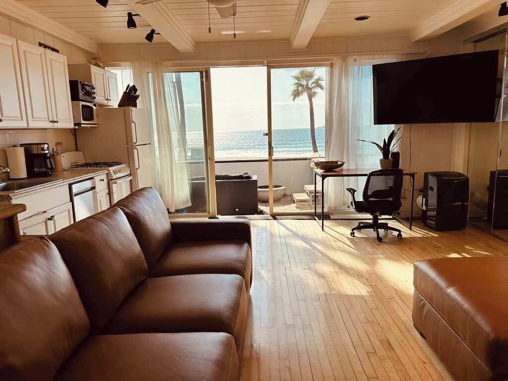 A modern living room with a brown leather sofa, a kitchen area with white cabinets, a flat-screen TV mounted on the wall, and a large sliding glass door leading to a balcony with a view of the ocean.