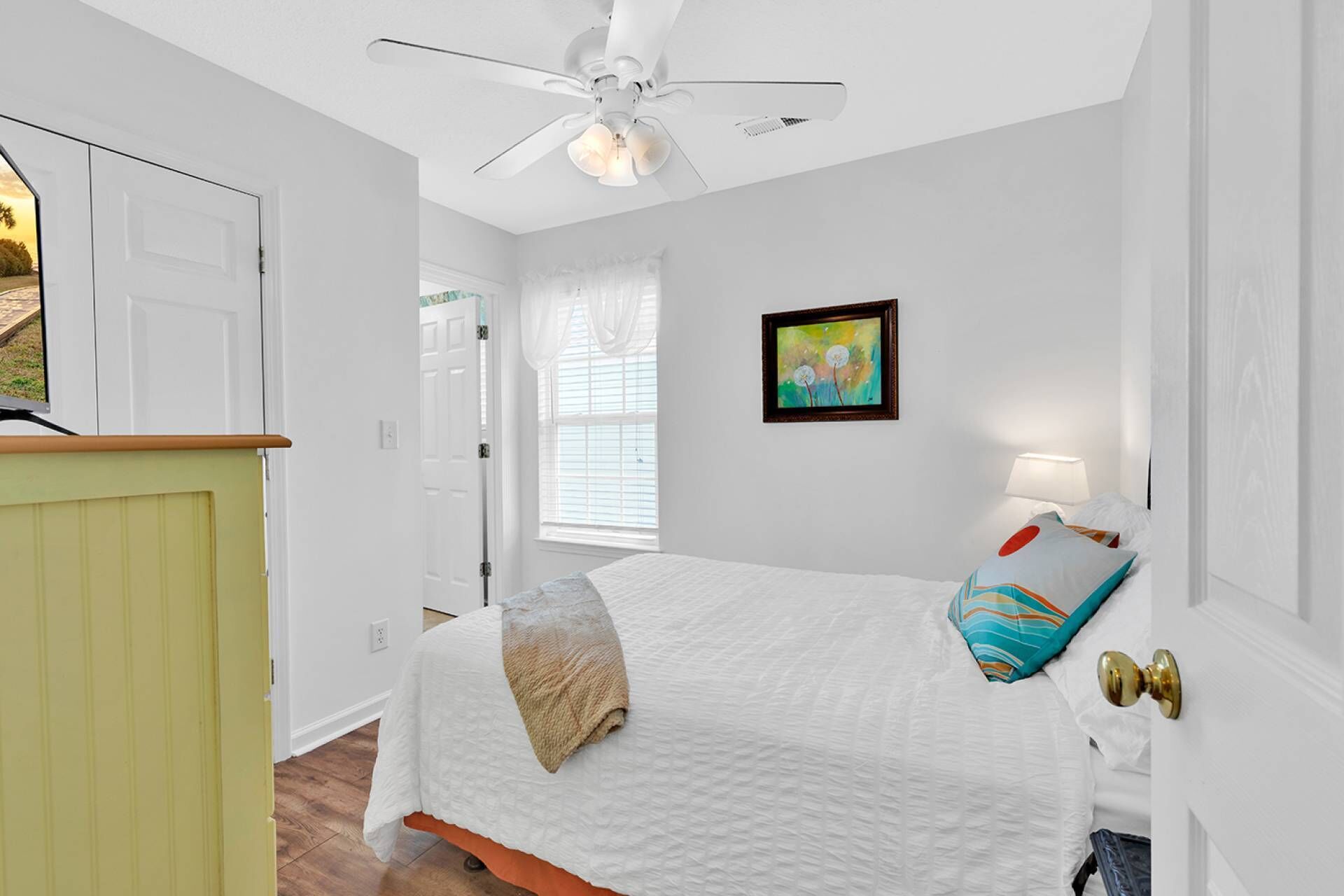 A bedroom with a bed, a ceiling fan, a window with blinds, a door, and a painting on the wall.