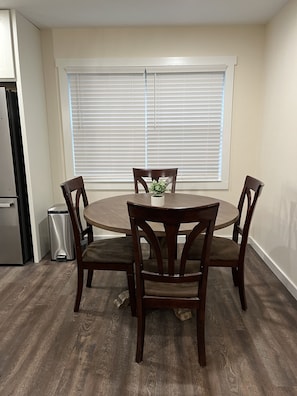 Un coin repas comprenant une table ronde et quatre chaises, une poubelle et une fenêtre avec des stores.